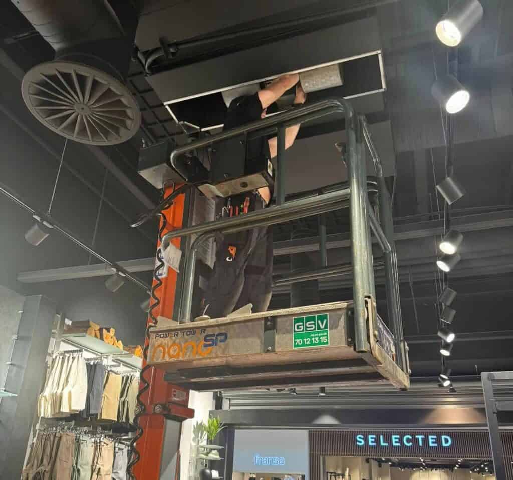 Montør arbejder fra lift på ventilationskanaler i loftet over en butik