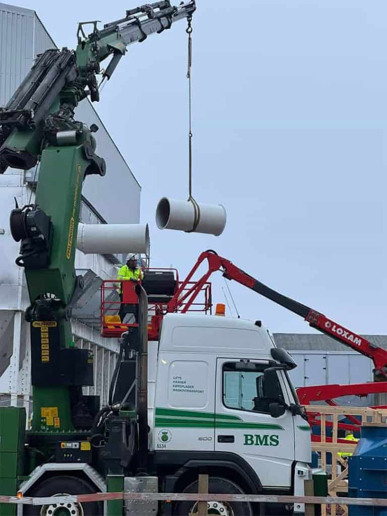 Lastbilkran løfter ventilationsrør på plads på et industriområde